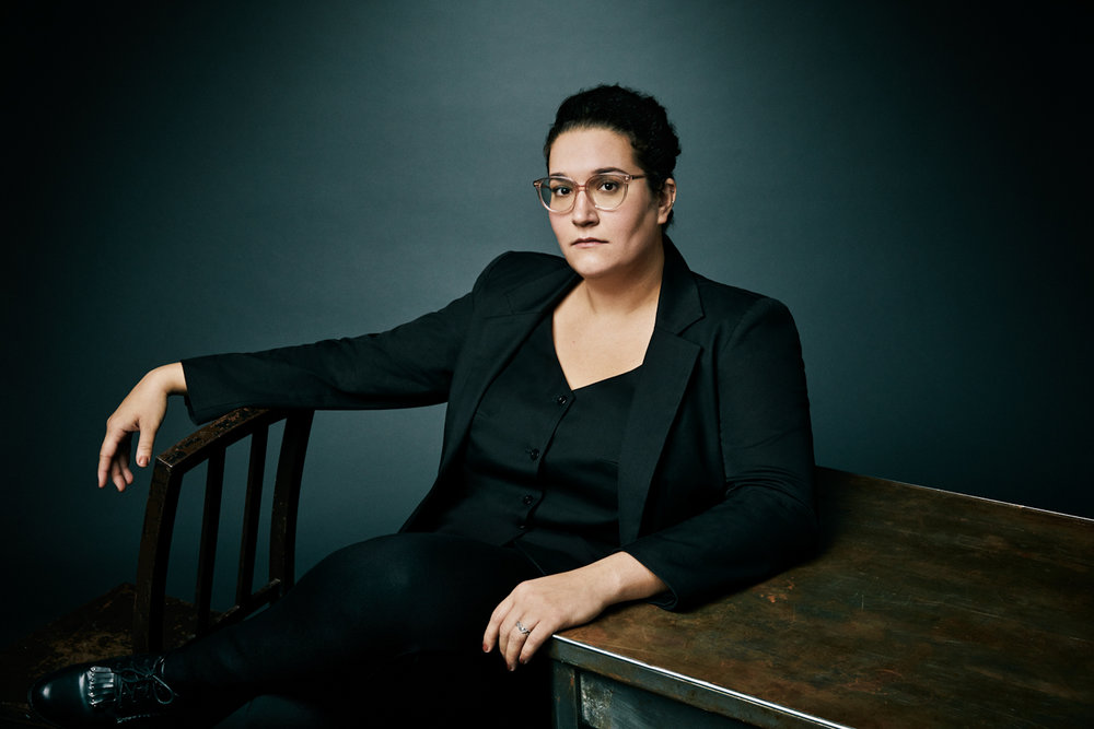 Carmen Maria Machado sits on a chair while leaning on a table on her right side. She is wearing a full black suit and black blouse. She has a stoic look as she stares into the camera. She has short dark hair and is wearing thin wireframe glasses